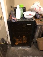 Front view of black Vissani wine fridge with glass door showing racks inside and some dust, located against a wall on a concrete floor with household clutter on top and nearby.