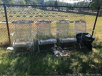 Four white metal patio chairs with mesh seating and backs, showing rust and wear, sitting outdoors on grass near chain link fence.