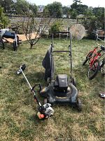 Full view of lawn mower and weed cutter placed on grass with surrounding yard items.