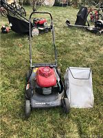 Full view of Honda Harmony HRT216 lawn mower with black metal body, red engine cover, black handlebar, and gray grass catcher bag placed next to it on grass.