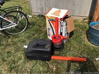 Wide view of the Black & Decker box beside the red blower with attached orange discharge tube, and the black Homelite case on grass.