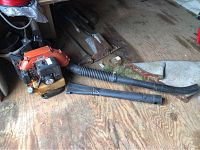 Photo showing the full backpack leaf blower unit laid on a wood floor with two black blower tubes extended and lying beside it, showing general condition and components.