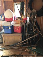 Photo showing stacked coolers in red, white, and blue, along with a pile of yard tools leaning against a wall, including rakes and shovels.