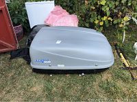 Full view of gray X-Cargo by Sears car top cargo carrier placed on grass near bushes and backyard items