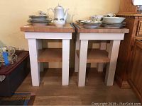 Pair of butcher block side tables with white legs and natural wood tops, stacked plates and teapot on one table.