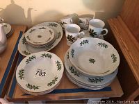 Set of porcelain dinnerware including plates, bowls, mugs arranged together on table. Holly leaf and berry painted decoration on white porcelain.