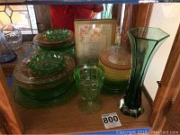 Display of green glass plates, ashtray, and bud vase with a framed print behind them on wooden shelf.
