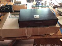 Closed hardwood silverware case with dark finish sitting on a cardboard box on a wooden table.