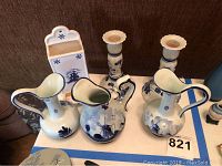 Photo showing 3 Delft-style ceramic pitchers, 2 matching Dutch ceramic candlesticks, and 1 repaired hanging match holder, all with blue and white floral and scenic decorations.