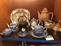 Photo showing group of four small blue and white porcelain teapots, miniature serving dishes, large wicker patterned ceramic teapot, Coalport oblong dish, black cast iron kettle, and other porcelain pieces arranged on shelf.