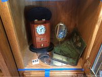 Wood mantel clock with storage drawers, tin match holder with floral design, ornate metal hair comb, small standing oval brass mirror, and green floral painting on board inside wooden cabinet