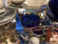 Shelving unit holding various glassware including blue glass bowls, clear glass bowls, pottery jugs, and plates showing arrangement and item variety