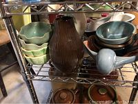 Metal shelving unit holding seven small green ceramic bowls, one large dark brown vase, and some blue-grey bowls. Ceramic pieces have earthy green and blue shades.