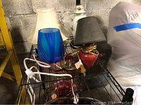 Photo showing two library lamps, two lamp shades, blue vase and clock on metal rack against brick wall.