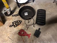 Photo of lot including a round cast iron skillet, corn bread pan, two trivets, a circular trivet stand, and a red-painted cast iron lobster decorative item.