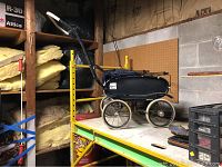 Side view of the vintage baby carriage showing its frame, wheels, and fabric body on a metal shelf.