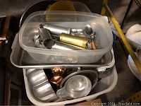 Plastic container with metal kitchen utensils, scoops, and cake decorating tools, resting inside a larger container holding baking tins and molds