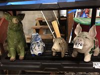 Four pig figurines on black shelving including green ceramic pig, blue and white porcelain pig, beige porcelain pig, and painted carved wood pig.