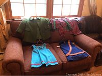 Green children's textured coats, blue sleeveless child's dress, blue skirt, and red and green plaid children's clothing laid out on a brown couch