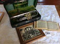 Photo showing antique silver-tone trumpet inside green-lined case, framed black and white military band photo, and open Christian hymn book on a table