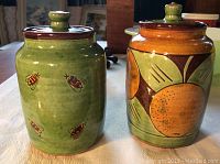 Front view of two pottery pots on white tablecloth, green honey pot with bees on left, marmalade pot with orange fruit design on right.