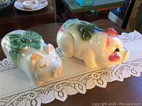 Two cream ceramic piggy banks with green bows and pink facial details, set on lace table runner