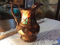 Front left side angle of the copper luster pitcher showing the handle, spout, and decorative motif including green leaves and gold accents.