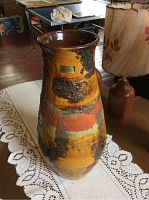 Full view of the Carstens lava glaze vase on a table with lace cloth, showing its tapering silhouette and textured glaze in warm earthy colors.