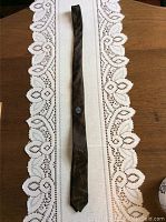 Full length photo of the dark colored tie lying on a white lace tablecloth, showing all printed signatures and a circular logo patch.