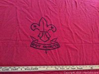 Close-up showing the dark Boy Scouts emblem with fleur-de-lis and a banner on red wool fabric.