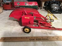 Side view of vintage Tru-Scale metal farm harvester showing red painted metal body, wheels, chain mechanism, and front hitch, ruler underneath for size reference approximately 12 inches long.