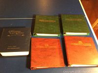 Five binders arranged on table: two green, two red, and one blue binder with gold embossed lettering showing titles of various stamp collections and first day covers.