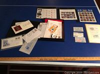 Overview photo showing mixed new and used stamps, first day covers, album pages and stamp sheets on blue table with ruler.