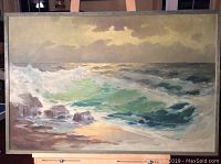 Full view of the ocean waves painting in a gold frame on an easel, showing the entire artwork with water, waves, rocks, and sky.