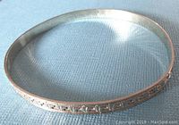 Top-down view of a sterling silver bangle bracelet with Greek key design, showing the entire round form and surface detail on a blue fabric background.