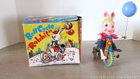 Photo of the Bell Cycle Rabbit wind-up toy with attached blue balloon and original box showing front and side graphics.