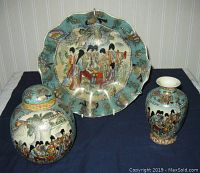 Three pieces of Asian pottery placed on a cloth: a large wall plaque, a ginger jar with lid, and a vase. Each piece is decorated with hand painted geisha garden scenes and raised beading with gold accents.