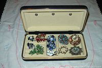 Photo of vintage rhinestone brooches and earrings in a cream-colored display case, featuring blue, green, white, amber, and turquoise stones.