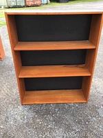 Front view of a teak wood bookcase with three shelves and black backing panel, showing the full height and width of the unit.