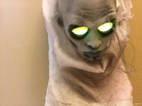 Close-up of ghoul's glowing eyes and face painted with dark eye sockets and blue lips in white fabric costume.