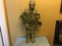 Full front view of the animated mummy Halloween decoration standing on a blue cloth with a neutral wall behind, showing its full height and wrapped bandage appearance.