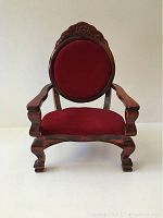 Front view of the vintage doll's chair showing the red velvet seat and backrest, carved wooden frame and armrests.