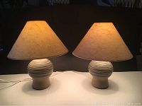 Pair of vintage pottery lamps lit, showing rounded striped bases and conical beige fabric shades on white tabletop against dark background.