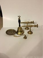 Two brass wall mount candle holders, vintage brass school bell with black handle, brass candle snuffer, and brass candle plate all arranged on white background.