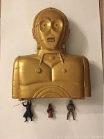Front view of the C3PO gold plastic carrying case with three Star Wars figures (Darth Vader, Jawa, and silver protocol droid) placed below.