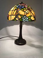 Full view of Tiffany style lamp with stained glass shade lit showing colorful dragonfly and floral patterns and dark metal base.