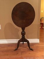 Front view of the vintage dark wood tilt top side table, top in vertical position, showing round shaped top and decorative curved legs.