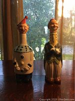 Two colorful vintage porcelain liquor decanters shaped as characters, standing side by side on a wood surface with a background of window and curtains.