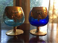 Photo of two Murano glass brandy snifters with hand painted floral and gold gilt decoration, placed on wooden surface with window background.