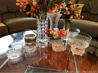 Glass table displaying multiple crystal bowls, vases, and faux flowers in various sizes and styles on a brown carpet background.